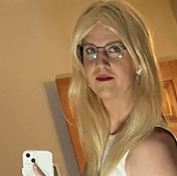 The TPERRY256 mascot photo of Todd, wearing white with glasses, full makeup, and blonde hair, taking a selfie in front of a wooden door